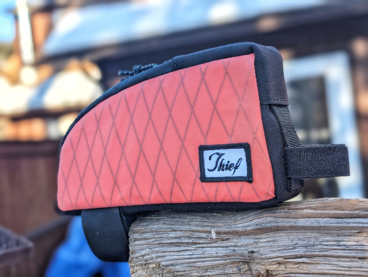 Thief Bikepacking Snack Break coral Xpac top tube bag with black trim and branded patch on a wooden post