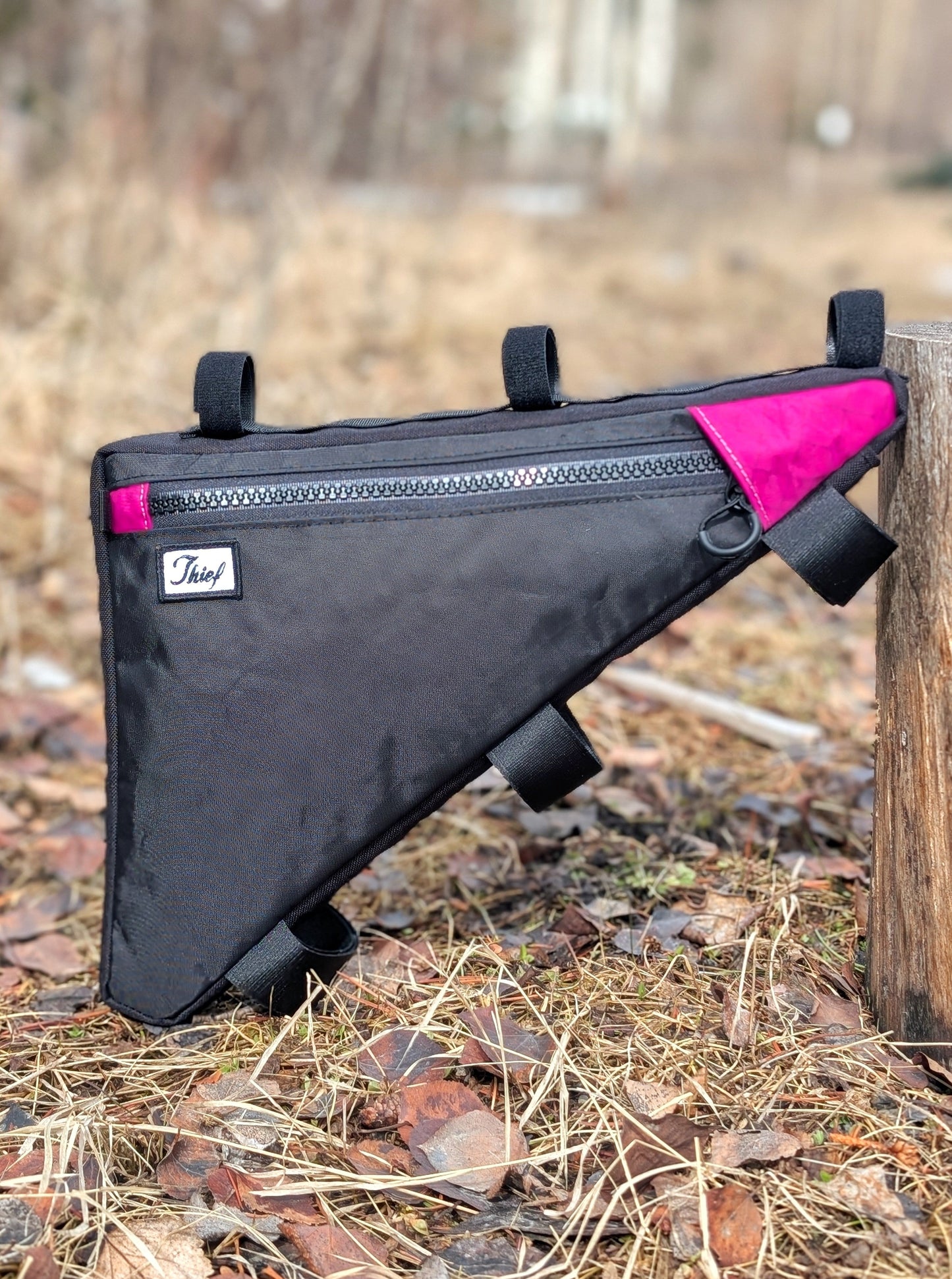 Black bike frame bag with pink accents attached to a wooden post outdoors.