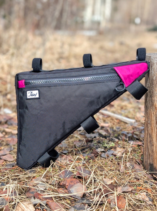 Black bike frame bag with pink accents attached to a wooden post outdoors.