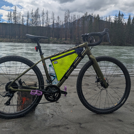An olive green gravel bicycle with a bright yellow frame bag is parked on a concrete riverbank with a river, forest, and cloudy sky in the background.