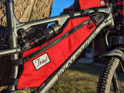 Red bicycle frame bag with 'Thief' branding attached to a bike, against a natural background.