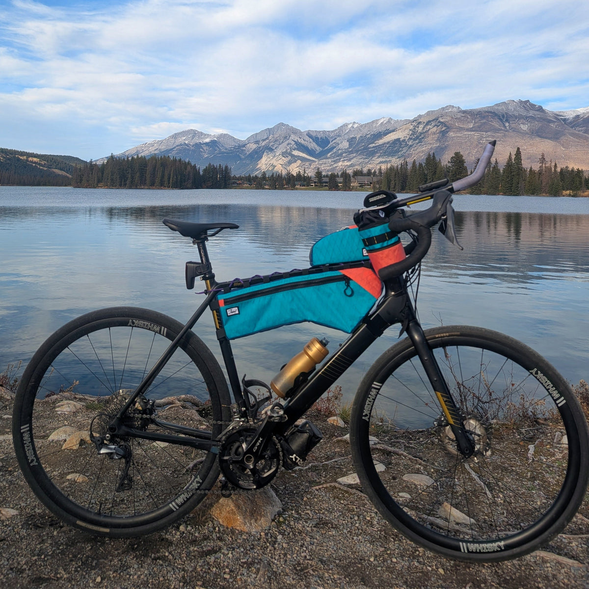 A black bicycle with a turquoise and orange top tube bag stands on a rocky lakeshore with mountains and blue sky in the background.