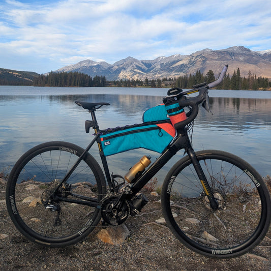 A black bicycle with a turquoise and orange top tube bag stands on a rocky lakeshore with mountains and blue sky in the background.
